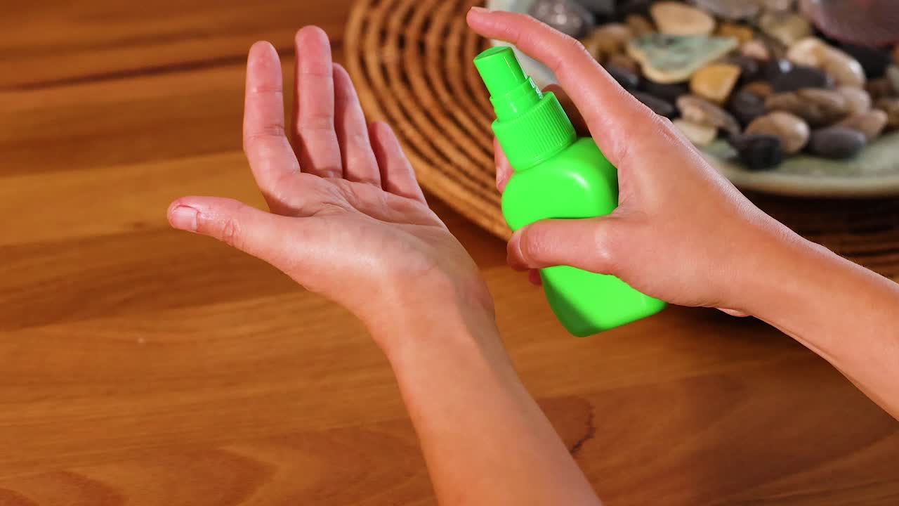 Hands applying sanitizer from a green bottle on a wooden table with decorative stones in the background
