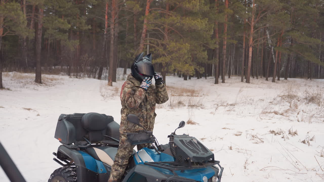 Il guidatore ispeziona il quad nella neve vicino ai pini, camminando intorno al quad blu controllando pneumatico, verricello e motore, regolando casco e cinghie, manutenzione metodica prima di un'escursione fuoristrada estrema.