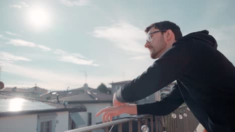 young attractive caucasian man with beard and glasses on balcony, sunshine pointing at happening in distance, watching people below him