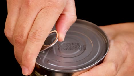 a human hand opens a metal canned food container using a pull tab, shown in close-up with sharp lighting and a black background