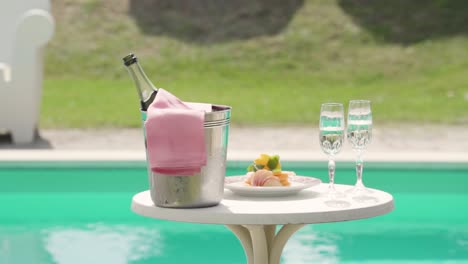 Botella-De-Champán-En-Un-Cubo-De-Hielo-Con-Un-Par-De-Copas-De-Vino-Y-Comida-Gourmet,-Piscina-Spa-Hotel-Resort-En-El-Fondo