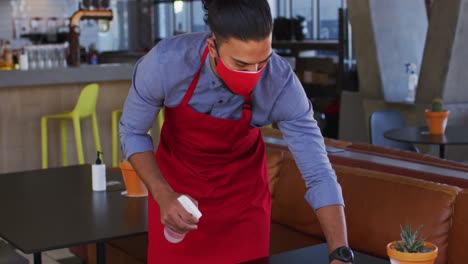 mixed race male cafe worker wearing face mask disinfecting table