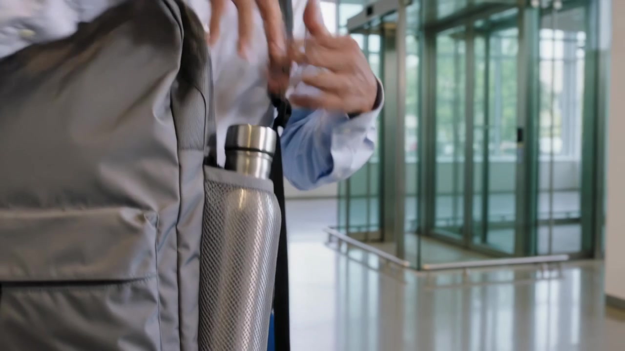Uomo in camicia blu che prende una bottiglia d'acciaio dalla tasca a rete di uno zaino grigio per bere nella hall.