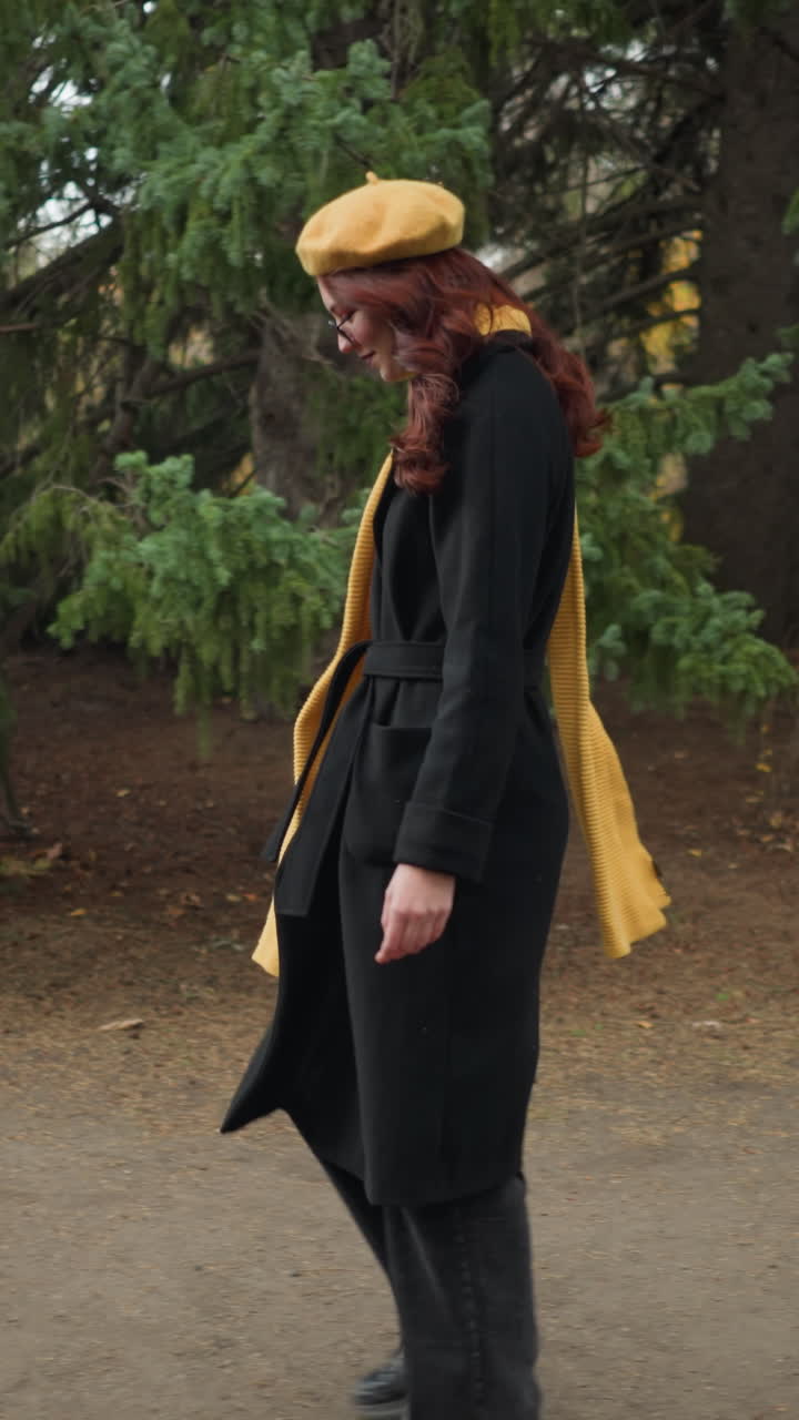 Turista felice con lunghi capelli rossi ondulati, che indossa un cappotto nero, un basco giallo e una sciarpa abbinata, mentre cammina attraverso un rigoglioso parco verde adornato da un colorato fogliame autunnale, irradiando gioia.