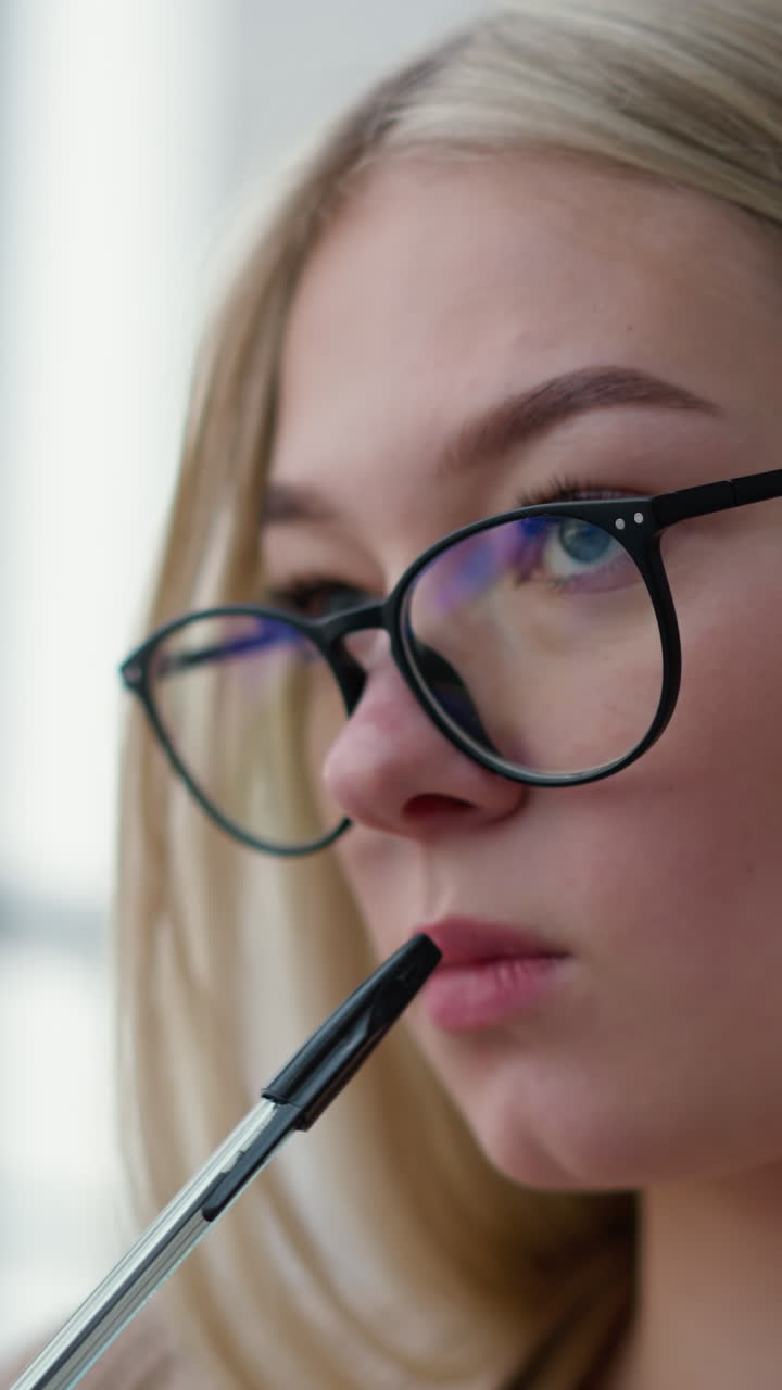 Close-up of pensive lady with glasses holding pen in her mouth, contemplating, then drops pen while gazing outside through a glass panel, reflecting a thoughtful and introspective moment