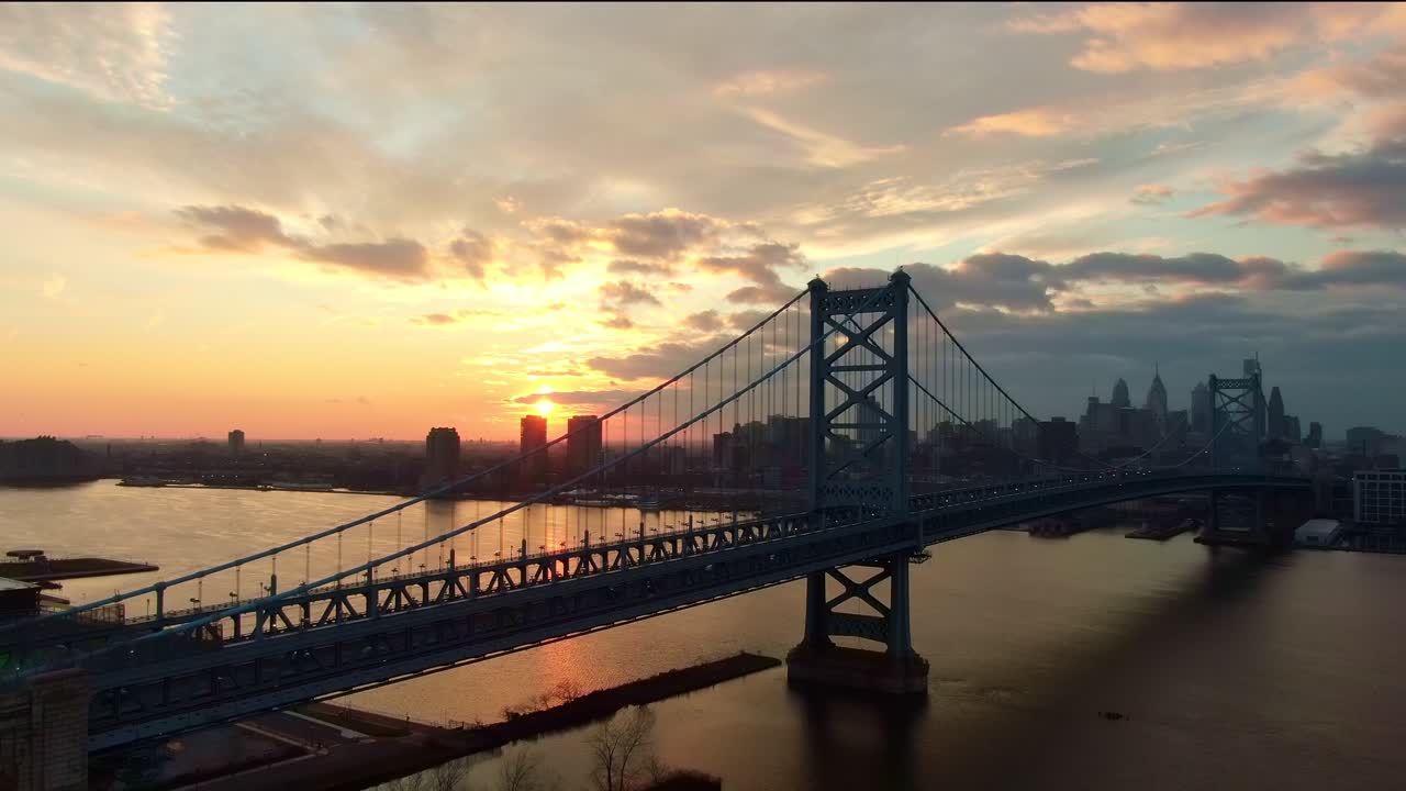 Ben Franklin bridge in dramatic sunset, aerial view of Philadelphia's famous landmark with skyline and modern skyscrapers in background, aerial pedestal view, establishing shot