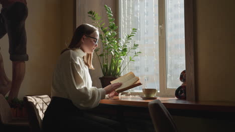 young student in white sweater reads novel near sunlit window, flipping to new page with focus and calm expression, surrounded by cozy indoor decor including potted plant and warm lighting