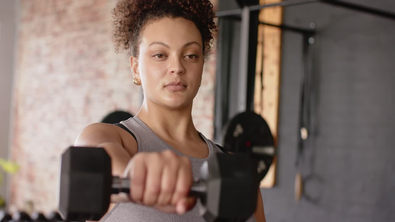 Woman gripping hexagonal dumbbell and lifting it forward inside gym with rig for shoulder workout