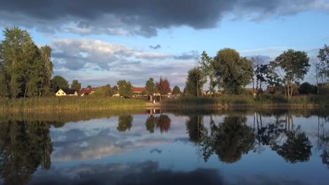 Vuelo-Aéreo-De-1-Millón-De-Dólares-De-Una-Chica-Rubia-Solitaria-Con-Vestido-Rosa-De-Verano-Que-Se-Refleja-Al-Atardecer-De-Verano-En-Un-Lago-En-Un-Pequeño-Pueblo-En-Brandeburgo,-Alemania