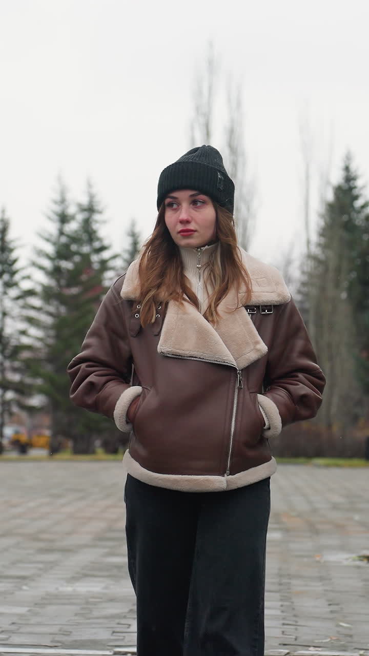 Portrait shot of young woman in black cap brown jacket walking slowly with hands in pockets on wide stone pathway through urban park lined with pine trees modern buildings and cloudy sky