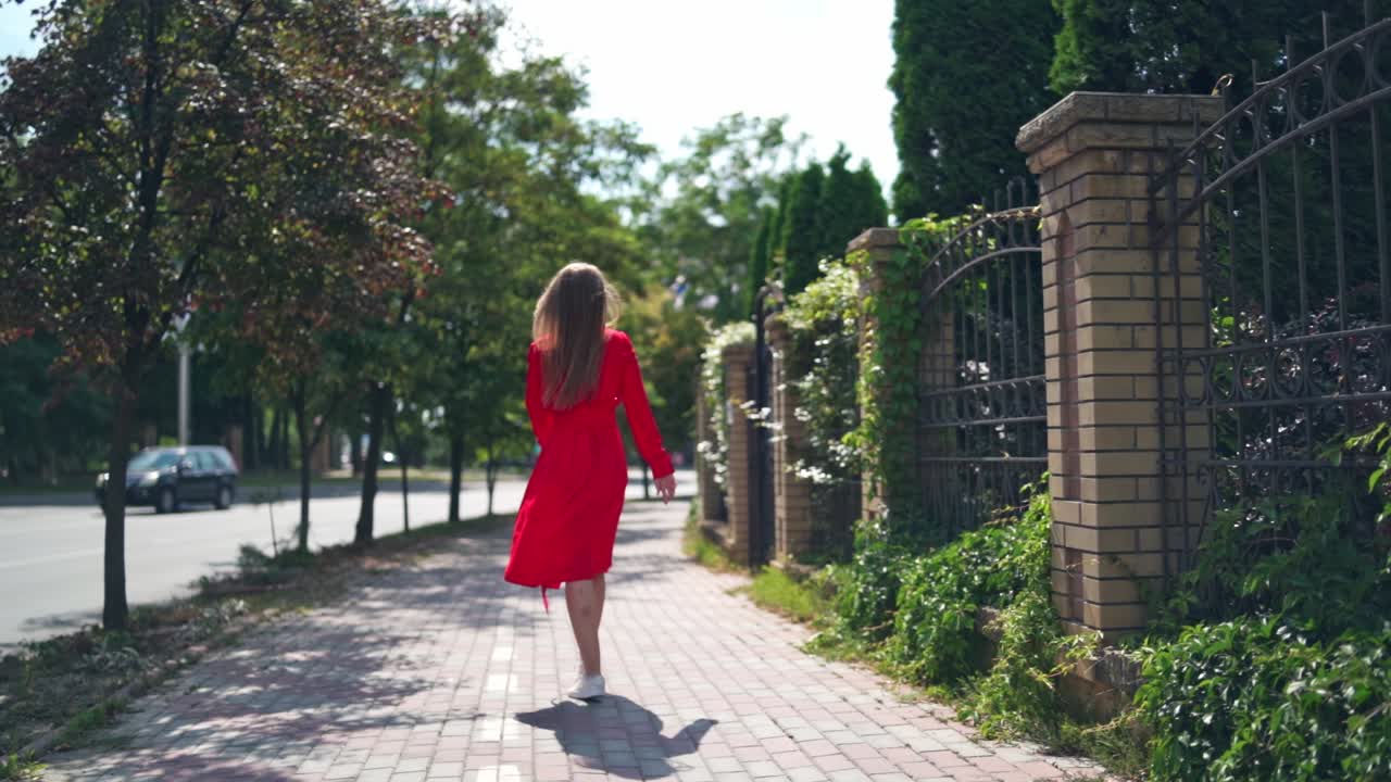 Backside view of a beautiful woman in the street. Young female in passionate red dress walking along the street and turning back happily.
