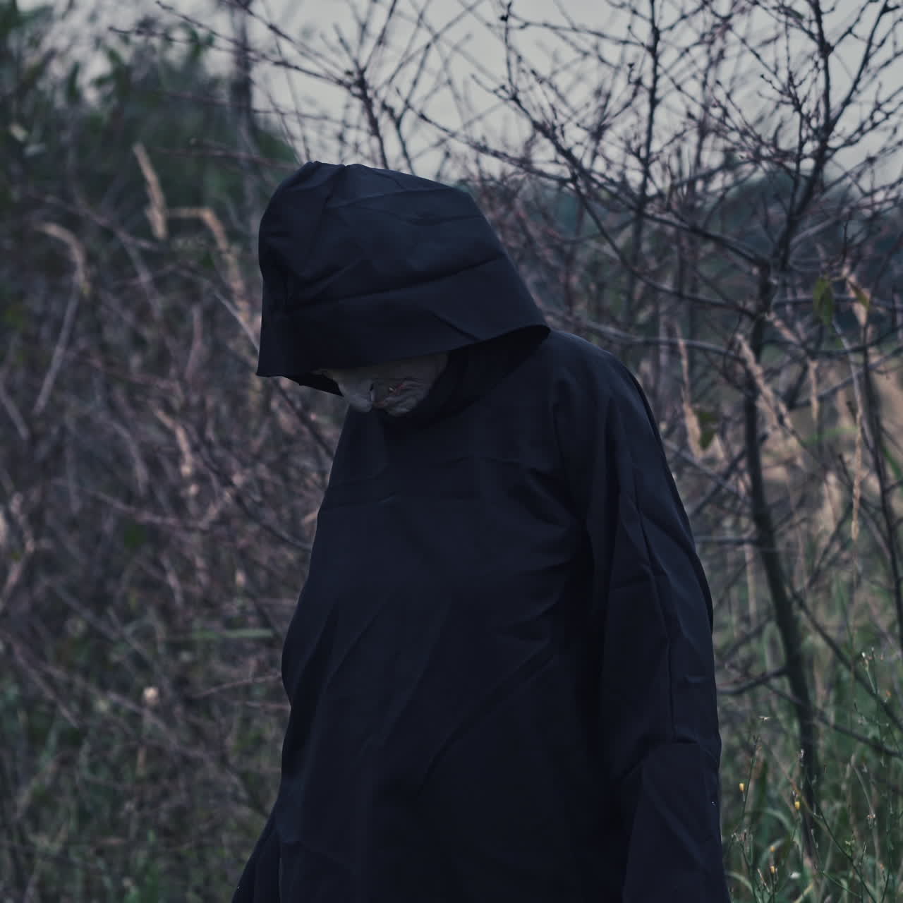 Horror figure in dark costume with hood. Portrait of scary ghost with outstratched arms on nature background. Halloween concept.