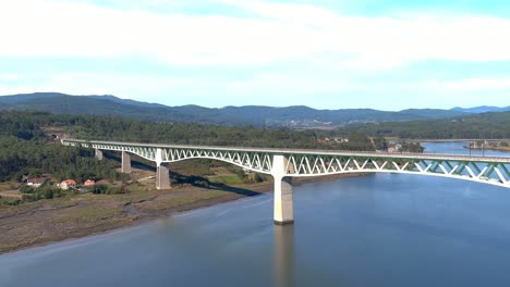 puente ferroviario sobre el río ulla, el pueblo, edificios industriales y la orilla, el soleado cielo azul orizonte, disparo de drones que viaja en diagonal hacia la izquierda, catoira, galicia, españa