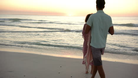 biracial couple stands close, wrapped in a pink blanket, watching a beach sunset with copy space