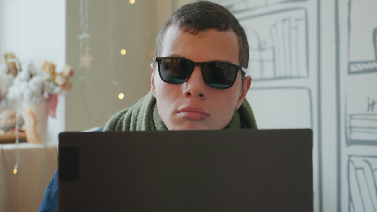 Kaukasischer Mann auf dem Sofa mit Laptop, Sonnenbrille und grünem Schal tragend, intensiv tippend, Nahaufnahme einer Innenaufnahme mit sanften Lichtern und illustrierter Bücherregal-Kulisse, gemütliche moderne Wohnung, Freiberufler.