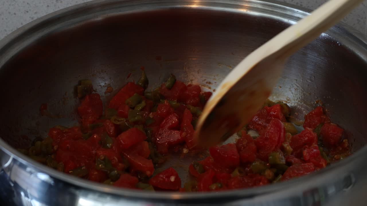 Stirring red stew tomato chunks in metal pot with ladle, close up of simmering dish preparation