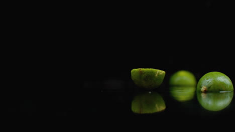 2-Reife-Limetten-Fallen-Auf-Ein-Glas-Mit-Wasserspritzern-In-Zeitlupe-Auf-Dunklem-Hintergrund