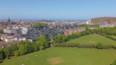 drone flies over green edinburgh meadows, cityscape to the left, and expansive holyrood park stretching in the background under clear skies