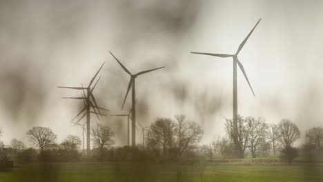 peaceful black and white depiction of a wind farm in a rural setting, featuring four in-motion three-bladed white wind turbines amidst trees and fields under a cloudy sky, with a foggy dream-like aura