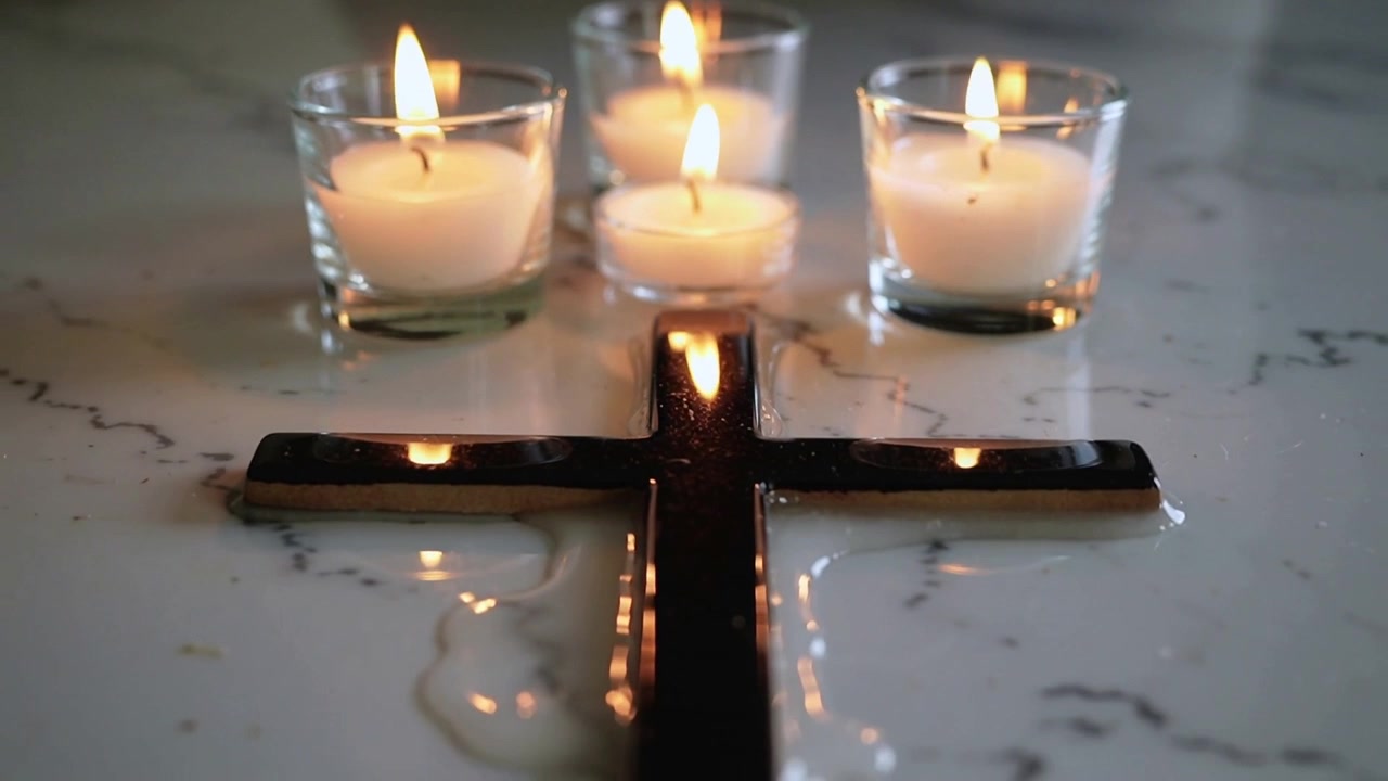 Tre fiammelle tremolanti di lumini in vetro sciolgono la cera in pozze su un piano di marmo, con una croce nera che si riflette.