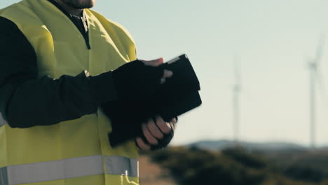 avanzando en energía limpia, un ingeniero profesional usa un chaleco reflectante mientras usa software de tecnología en una tableta para auditar turbinas eólicas en un día soleado en un campo de generadores de energía renovable