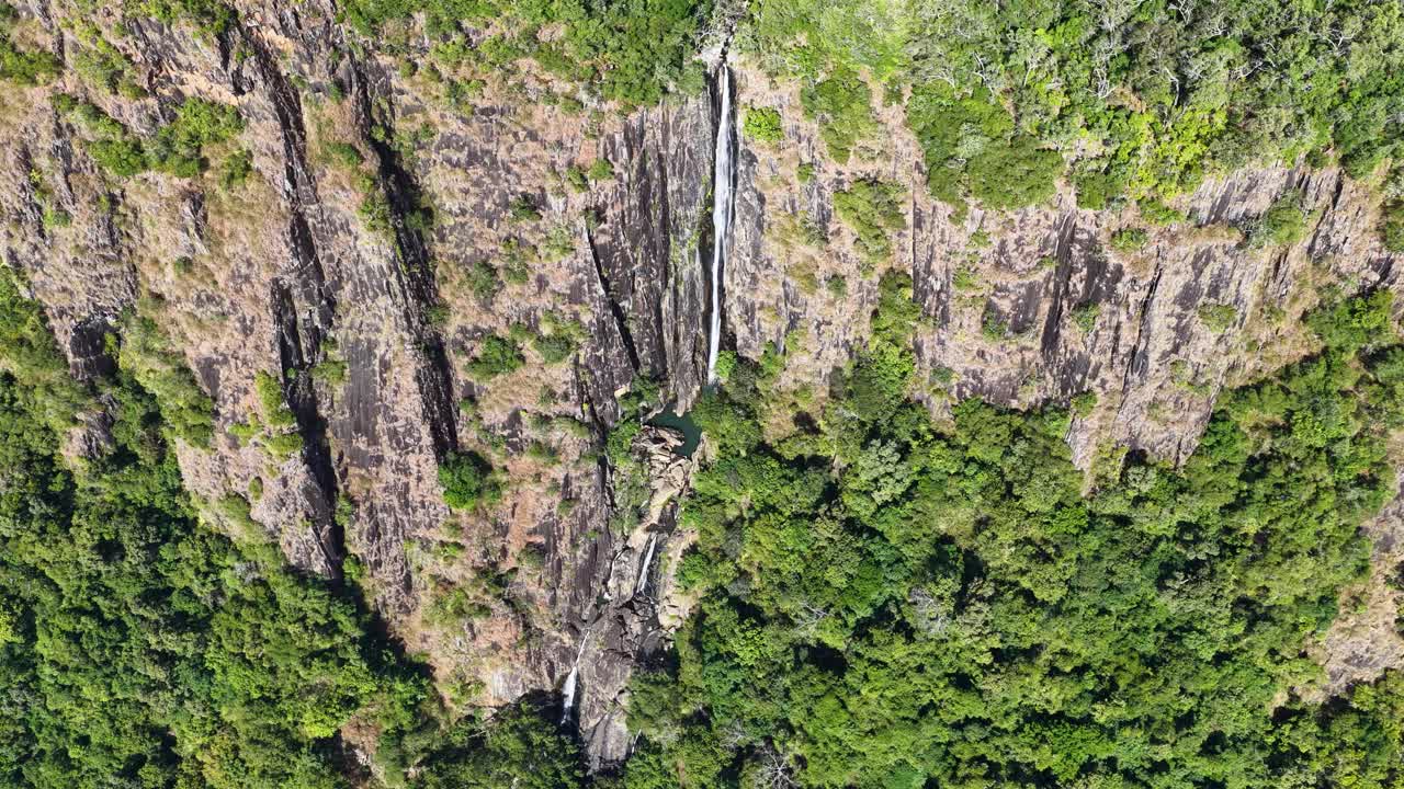 Drone footage captures a stunning waterfall cascading down rocky cliffs surrounded by lush greenery in the Daintree Rainforest