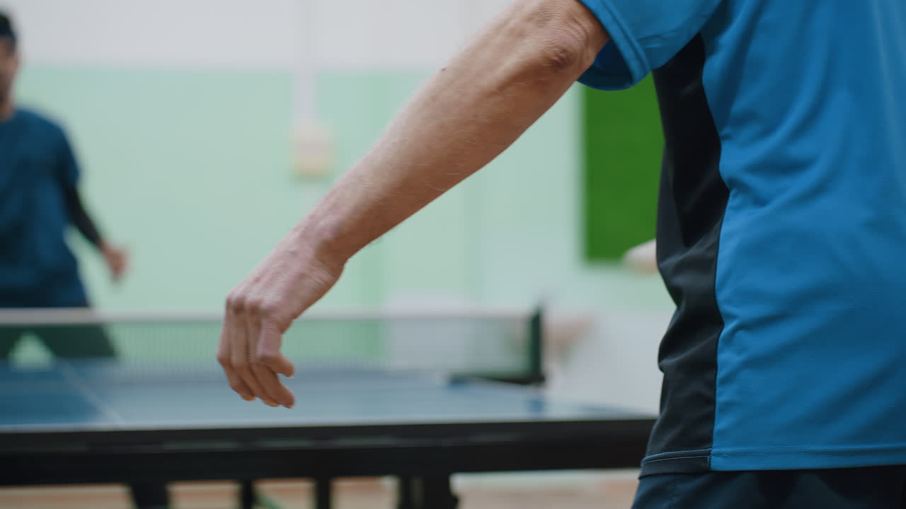 Close up back view of player in blue shirt during heated table tennis rally, opponent with paddle blurred in background, fast white ball visible mid-air showing energy of match