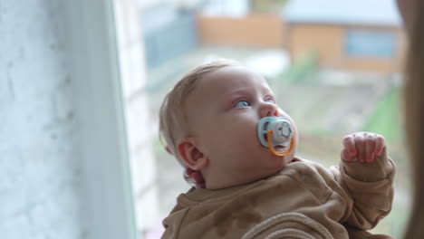 unrecognized woman waving her cute child near the window. calm baby boy with pacifier in mouth looks up.