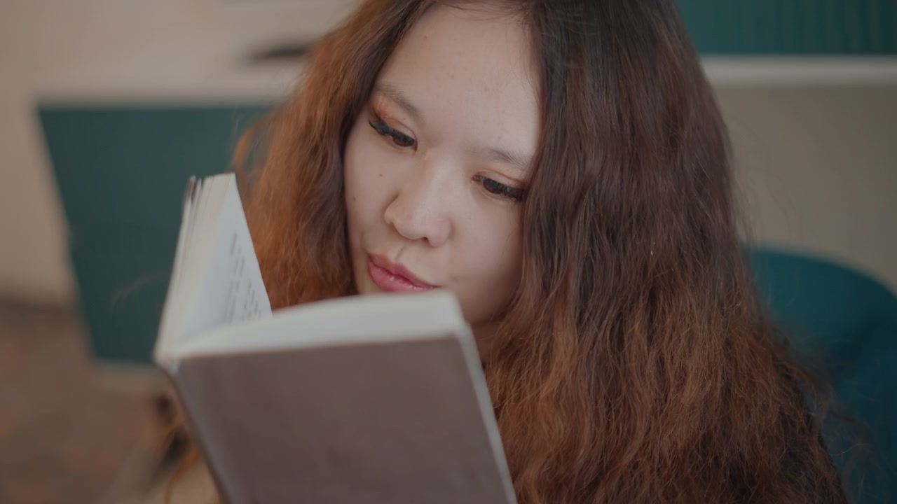 İç mekanda kitap okuyan genç bir kadının yakın görünümü; sabit aşağı doğru bakış, hafif baş eğimi ve nazikçe büzülmüş dudaklarla konsantrasyonu, sakin düşünceyi ve sessiz bir öğrenme ruh halini yansıtıyor.