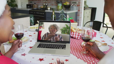 una pareja afroamericana sonriente con vino usando una computadora portátil para una videollamada de navidad con una mujer en la pantalla