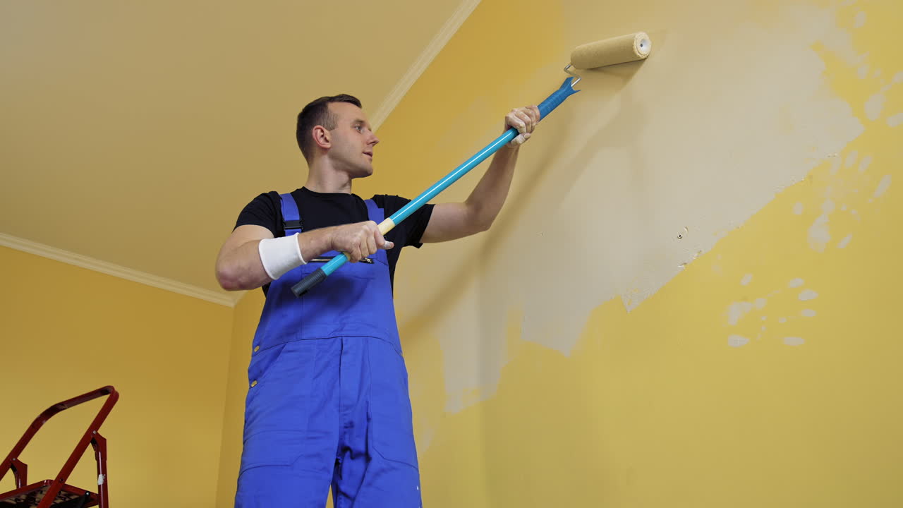 Builder painting wall in the room. Professional worker in workwear doing renovation in apartment with roller brush on a long stick. View from below.