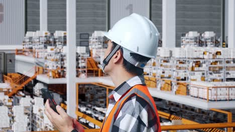 vista lateral de cerca de un ingeniero masculino asiático con casco de seguridad usando un teléfono inteligente y mirando a su alrededor mientras está de pie en el almacén con estantes llenos de productos de entrega