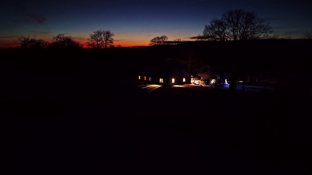Aerial rising shot reveals warmly lit cottages cars glowing against twilight blue hour sky silhouetted bare trees snowy field Latvia dusk landscape
