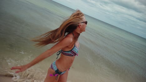 Closeup-slow-motion-of-laughing,-playful-mature-woman-in-bikini-and-sunglasses-turns-around-and-runs-on-the-beach