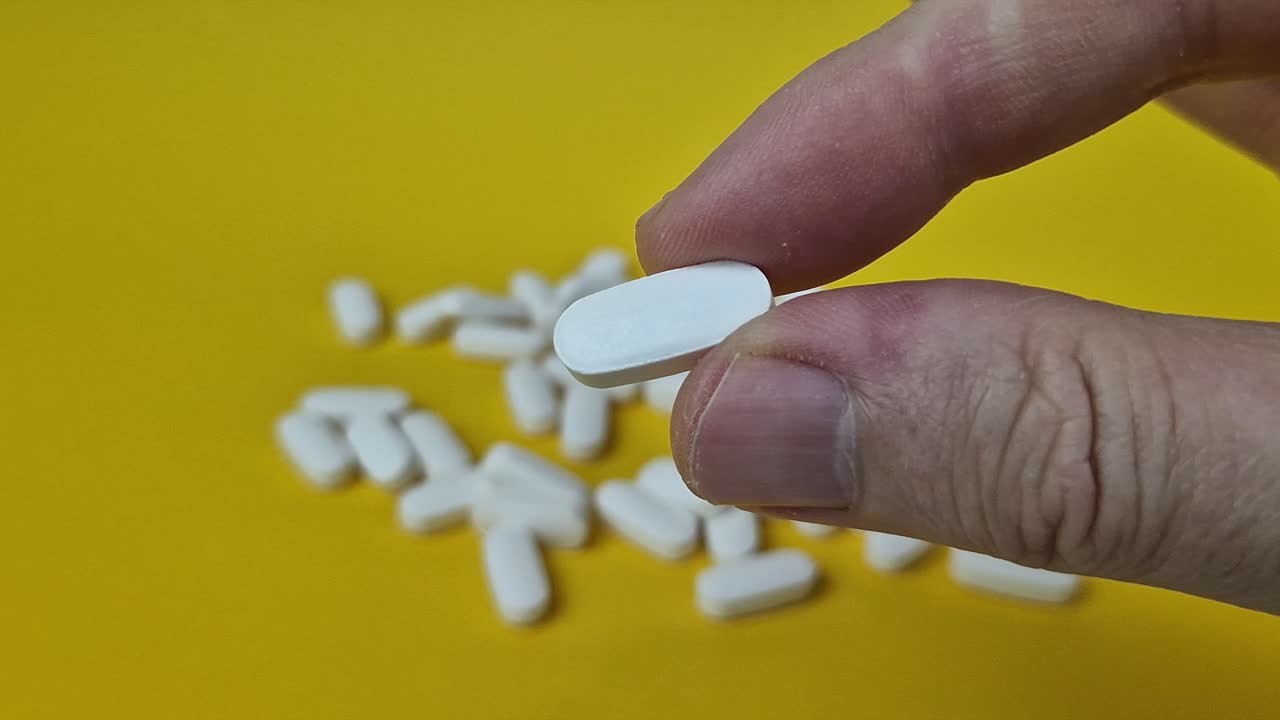 Extreme macro 4K close-up of a single white, oblong pill held delicately between a person's fingers against a bright yellow surface, with more pills blurred in the background
