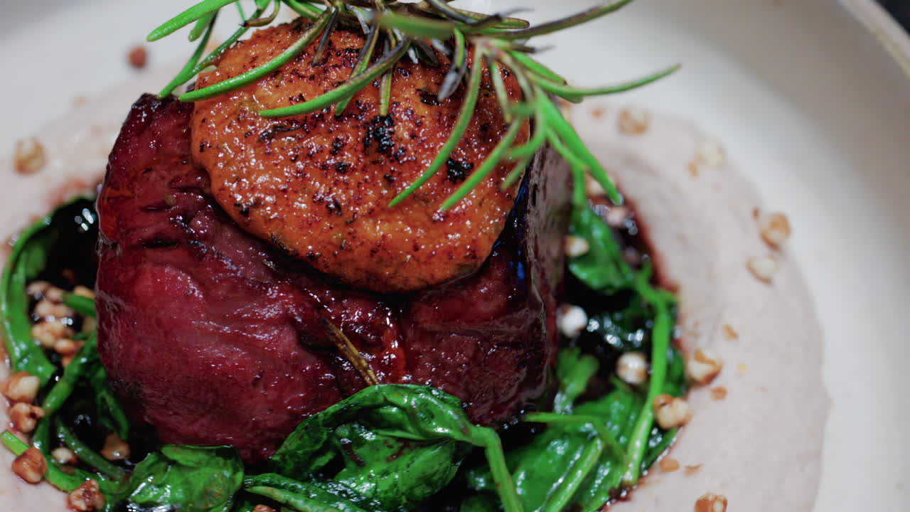 Close up of a gourmet dish featuring grilled steak topped with foie gras, served with sauteed spinach and a rosemary garnish