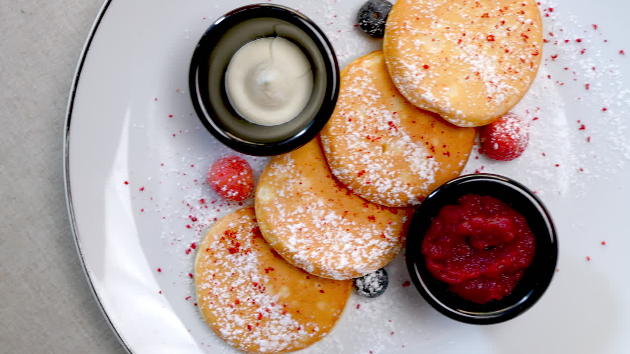 Small pancakes with powdered sugar, sour cream and berry jam are rotating on a white plate, creating a visually appealing presentation of a sweet treat