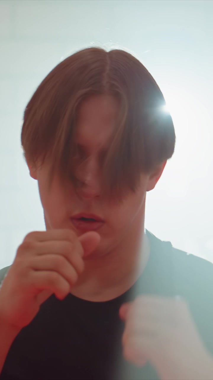 Young man with light brown hair wearing black shirt makes. Bright backlighting creates lens flare effects obscuring. Close up shots focus on torso up against plain background