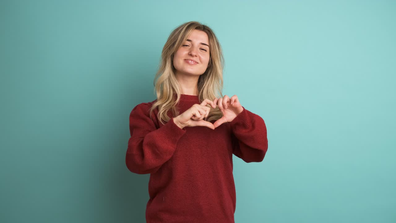 Happy woman blowing kiss and showing heart gesture in blue studio