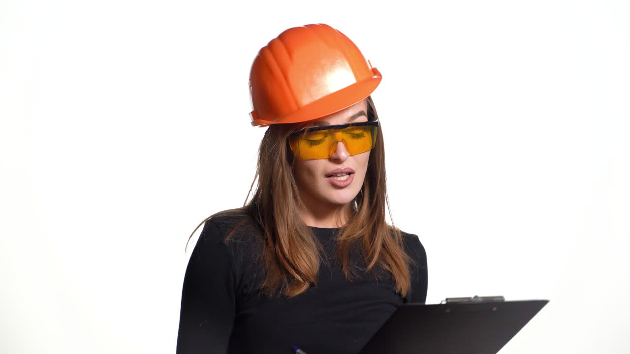An entertaining woman in orange glasses and hard hat is holding a folder and pen in her hands and talking with showing something on a white studio background.