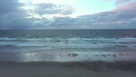 Aerial-view-away-from-a-person-walking-on-wet-reflecting-beach,-while-waves-hit-the-coast-of-North-Spain---Panning,-pull-back,-drone-shot