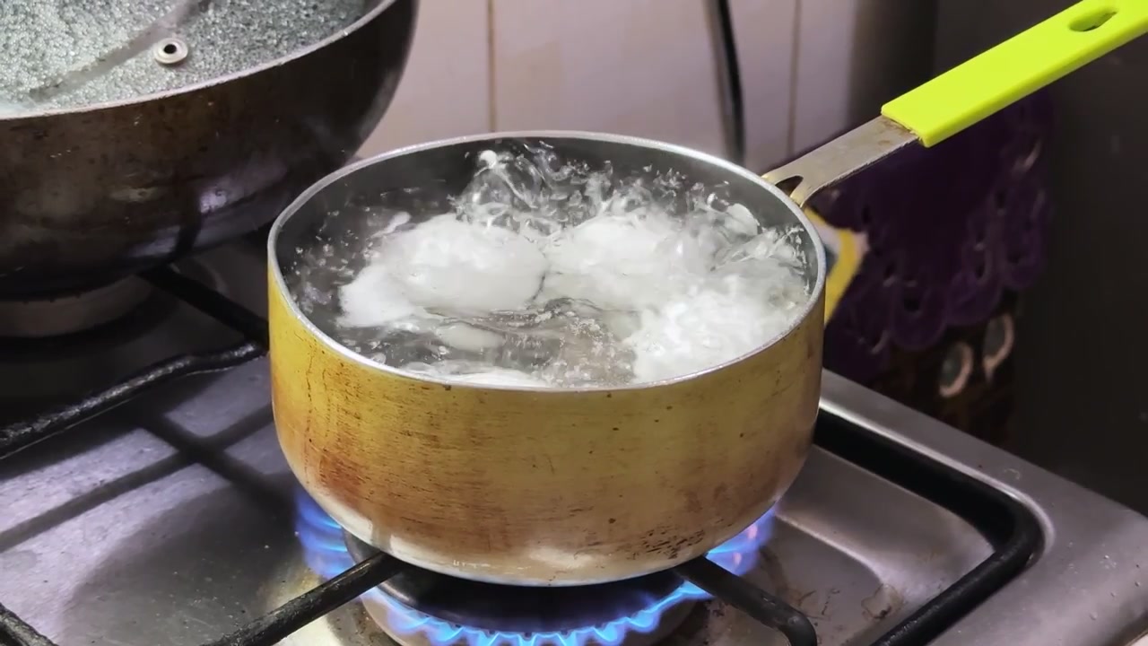 White eggs boiling in a small pot of water on a kitchen gas stove with blue flames. Bubbling hot water and steam during breakfast food preparation.