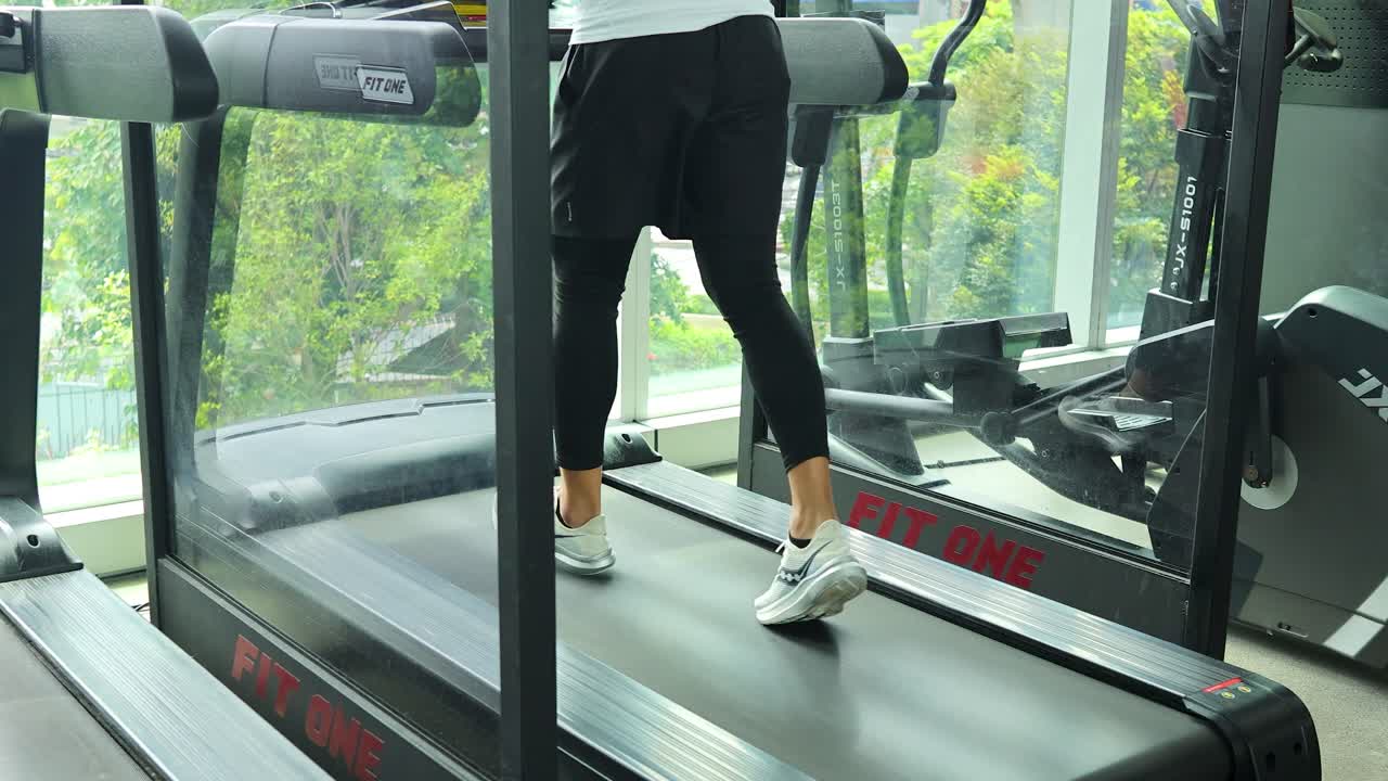 Person running on a treadmill in a bright modern gym with large windows and cityscapes