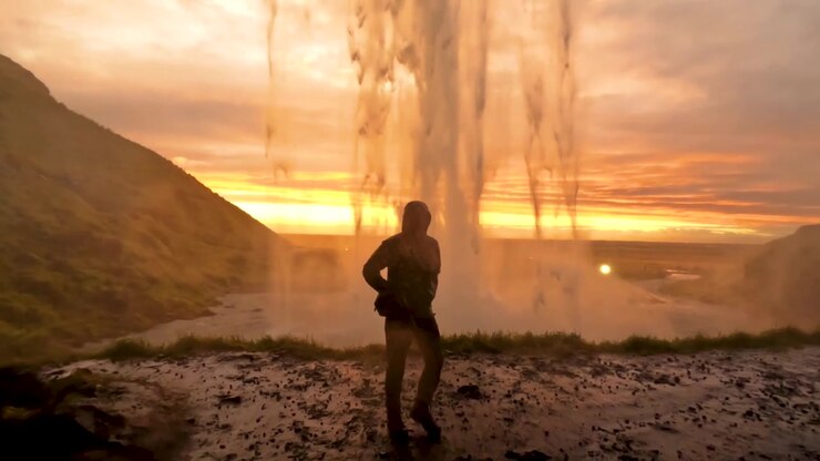 La telecamera si muove verso un uomo che si allontana sotto Seljalandsfoss al tramonto, paesaggio islandese.