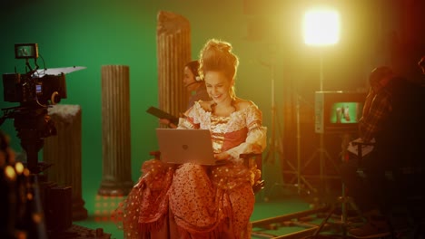 on period costume drama film set: beautiful smiling actress wearing renaissance dress, sitting on chair using laptop computer with green screen in background. studio shooting historical blockbuster