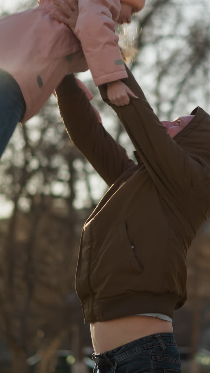 una niña con una gorra rosa y una chaqueta siendo levantada juguetón por un hombre con una chaqueta marrón y pantalones vaqueros azules. un momento alegre y cálido al aire libre con el hombre y el niño riendo