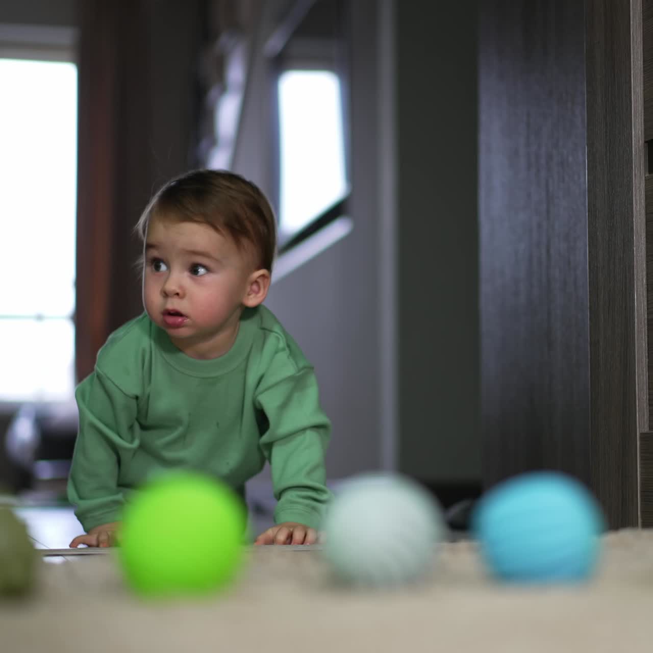 Adorable funny boy crawling by the floor approaching the camera. Lovely toddler looks at the various balls lying on the floor. Blurred backdrop