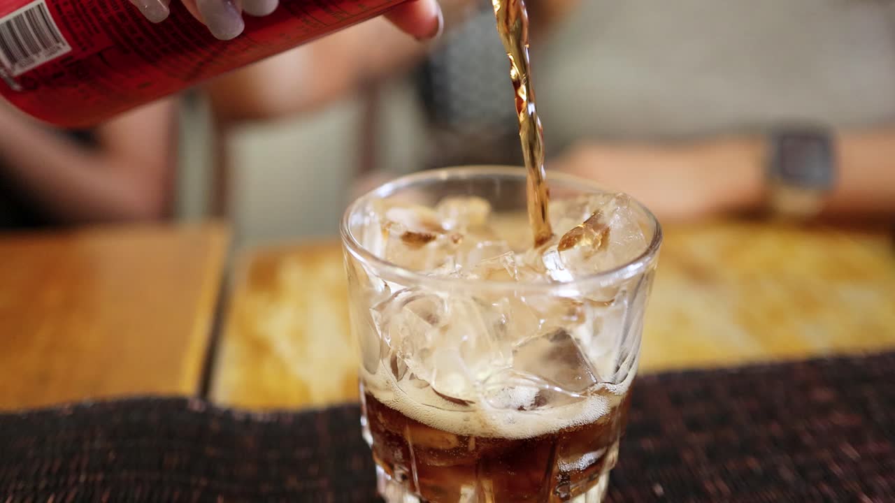 A close-up shot captures cola being poured into a glass filled with ice cubes on a wooden table, with soft indoor lighting and shallow depth of field