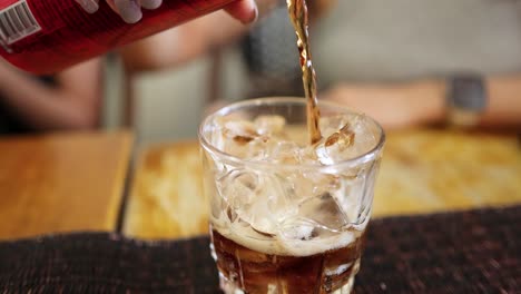 a close-up shot captures cola being poured into a glass filled with ice cubes on a wooden table, with soft indoor lighting and shallow depth of field
