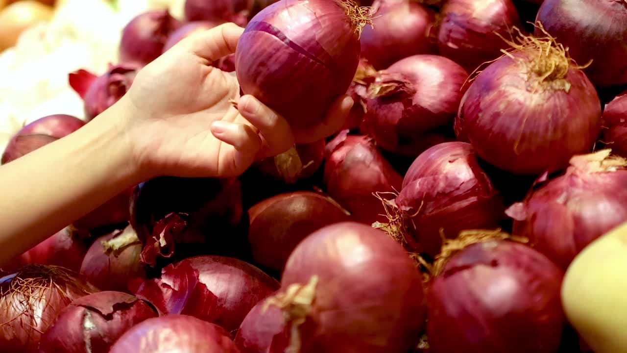 A hand carefully picks red onions from a vibrant, well-lit display in a grocery setting.